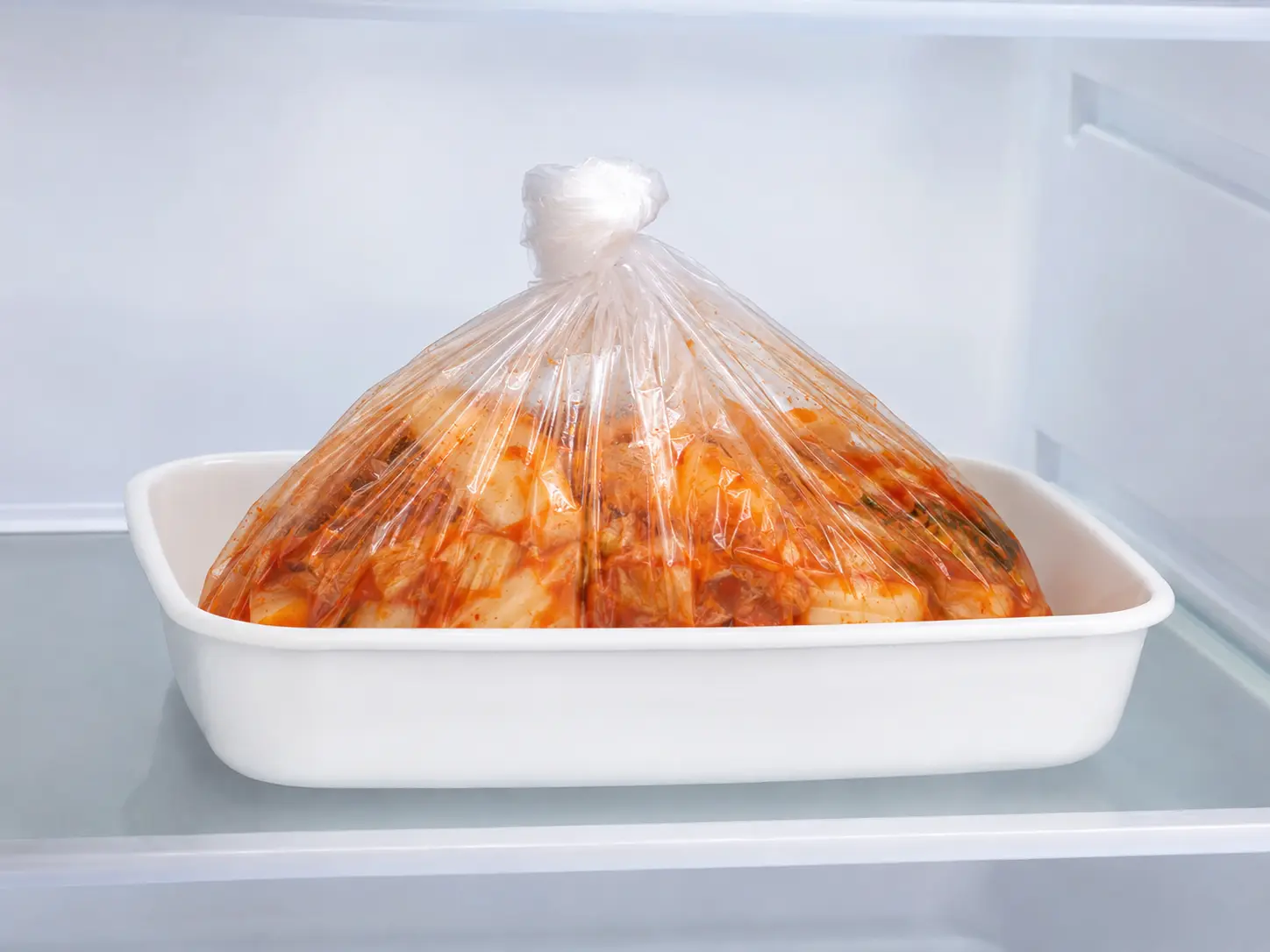 口を結んだ白菜キムチの食品用ポリ袋を清潔な容器へ入れ、整理された冷蔵庫で漬けている状態