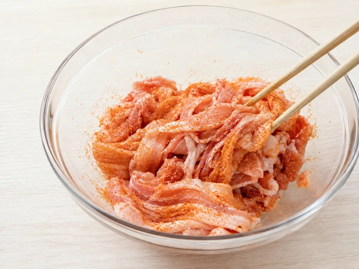 豚バラ薄切り肉へ赤橙色の粉末状のキムチ漬の素をまぶしている工程
