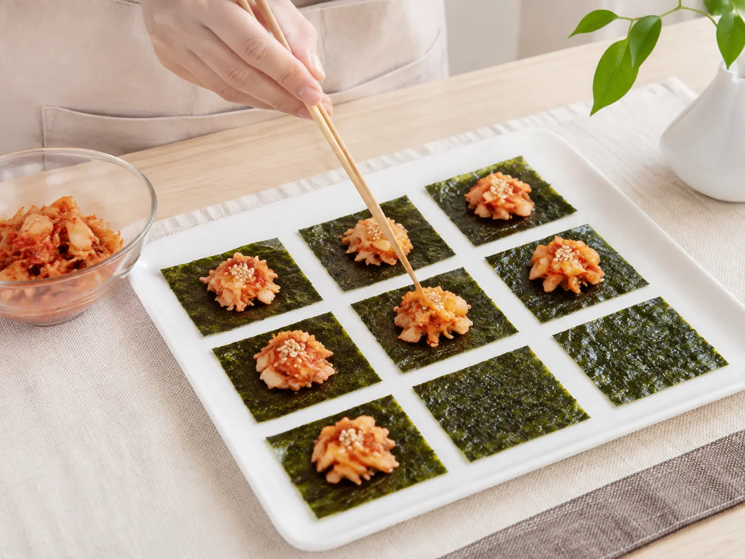 皿に並べた正方形の焼き海苔へ刻んだキムチをのせ、いりごまで仕上げる工程